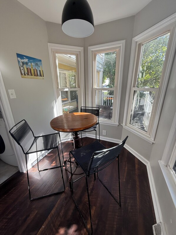 A dining area with a round wooden table, black metal chairs, and a pendant light. There are three large windows providing natural light and views of the outside.