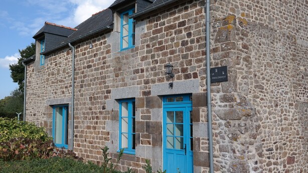 A stone building with blue windows and doors, a sign indicating a historical site, and a small garden area in front.