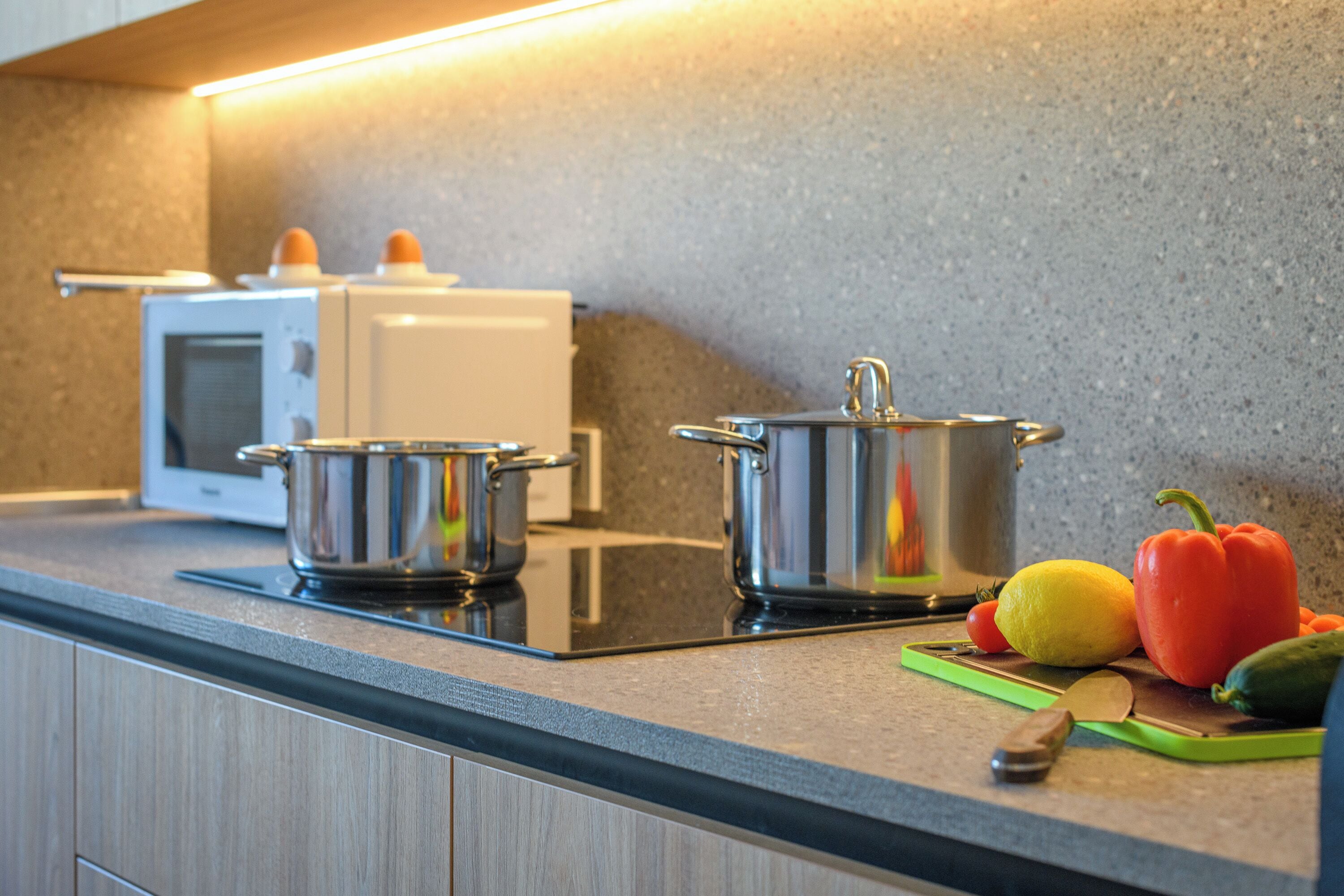 A kitchen with a microwave, two stainless steel pots, a cutting board with vegetables, and a knife.