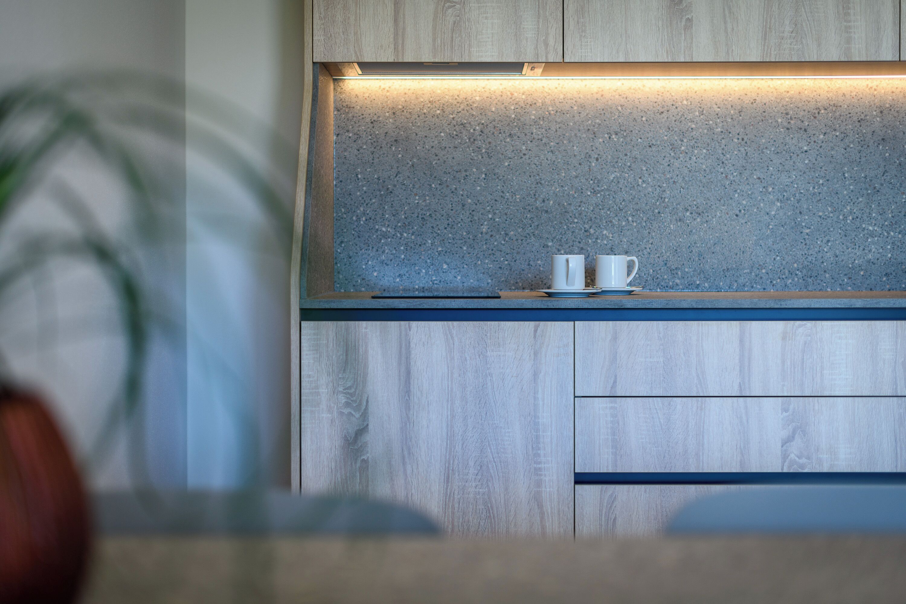 A kitchen with wooden cabinets, a speckled backsplash, and two white cups on a blue countertop.