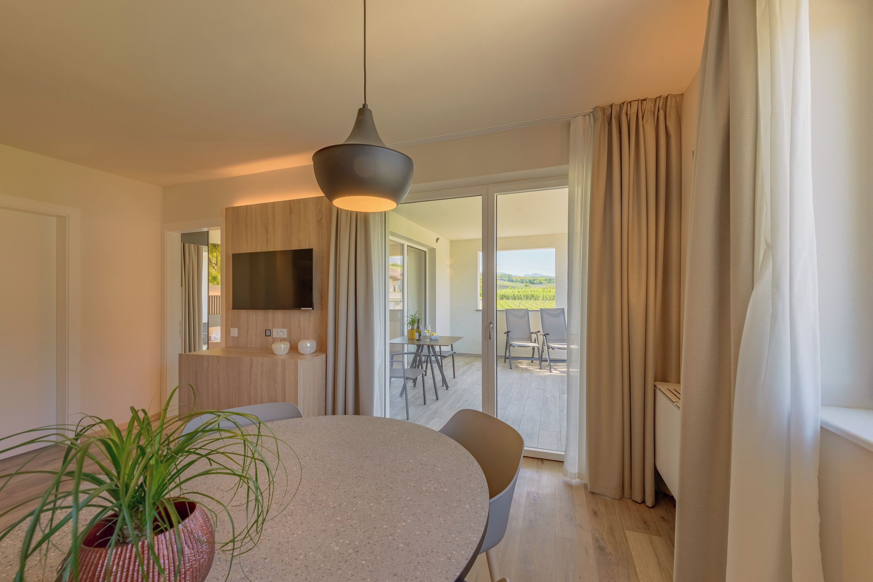 A modern hotel room with a round dining table, a potted plant, a flat-screen TV, and a sliding glass door leading to an outdoor patio.