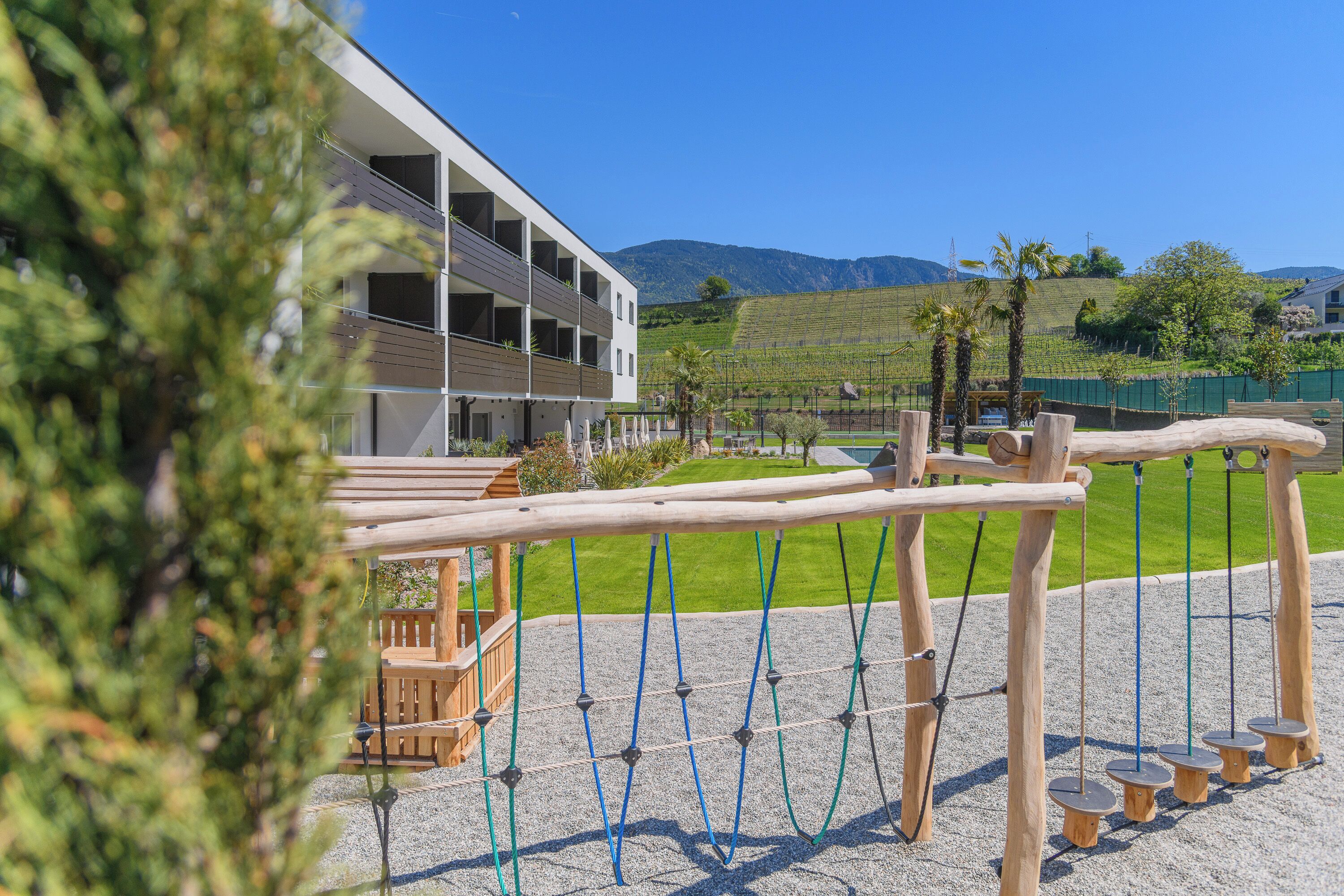 Un edificio moderno con un balcón, un parque infantil con columpios y estructuras para escalar, y un viñedo al fondo.