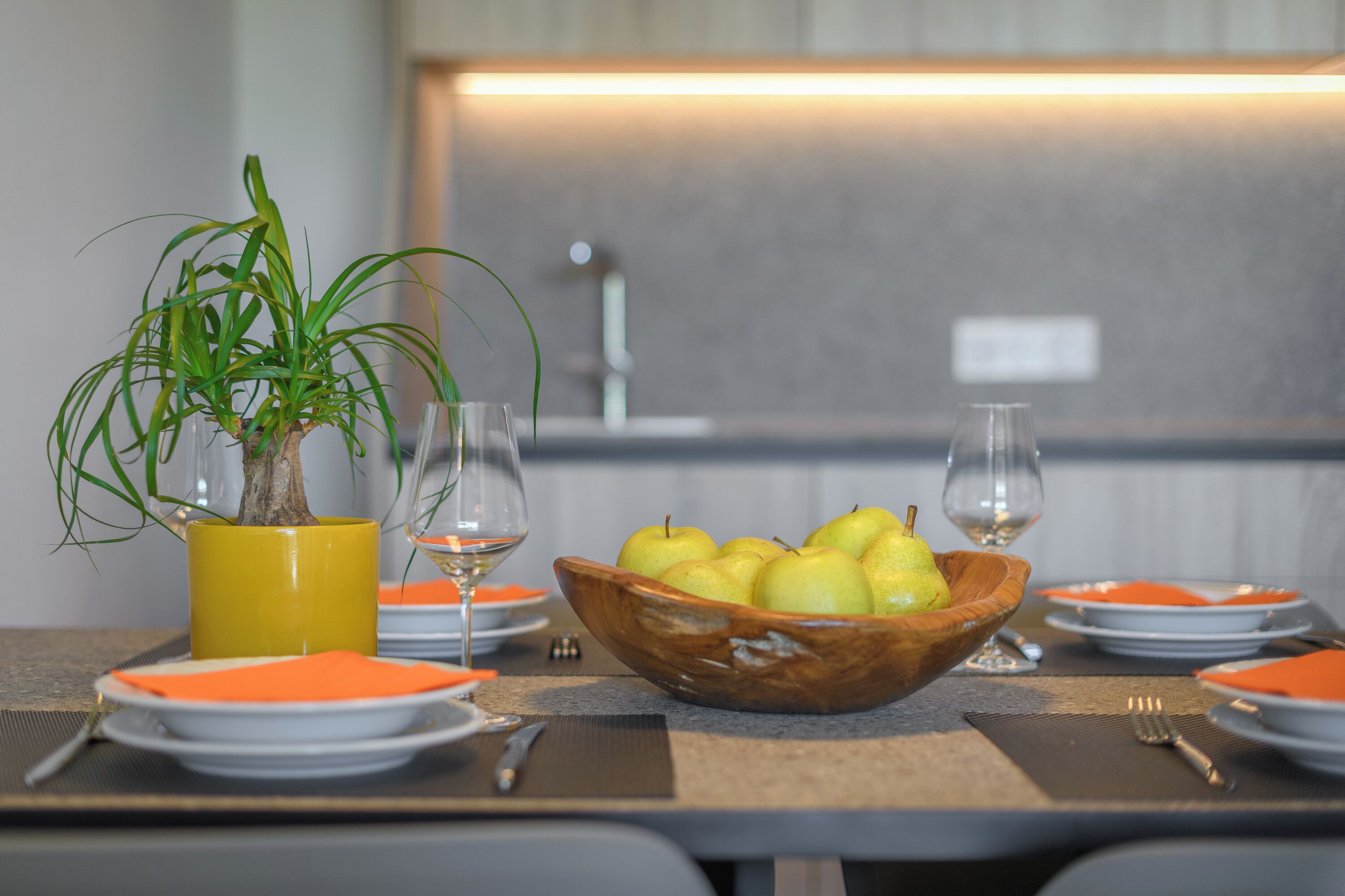 Una mesa de comedor con un cuenco de madera lleno de fruta, una planta en maceta y platos y cubiertos colocados cuidadosamente.