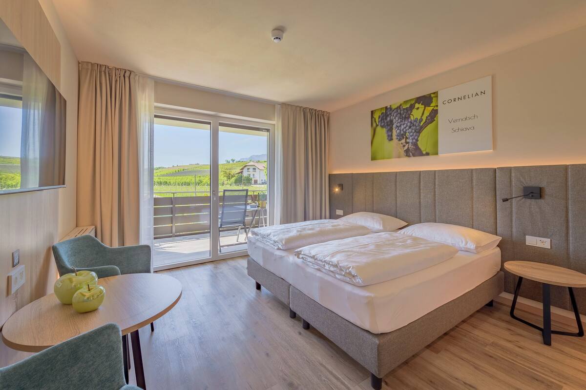 A hotel room with a bed, a table with chairs, a small round table, a sliding glass door to the balcony, and a wall-mounted artwork.