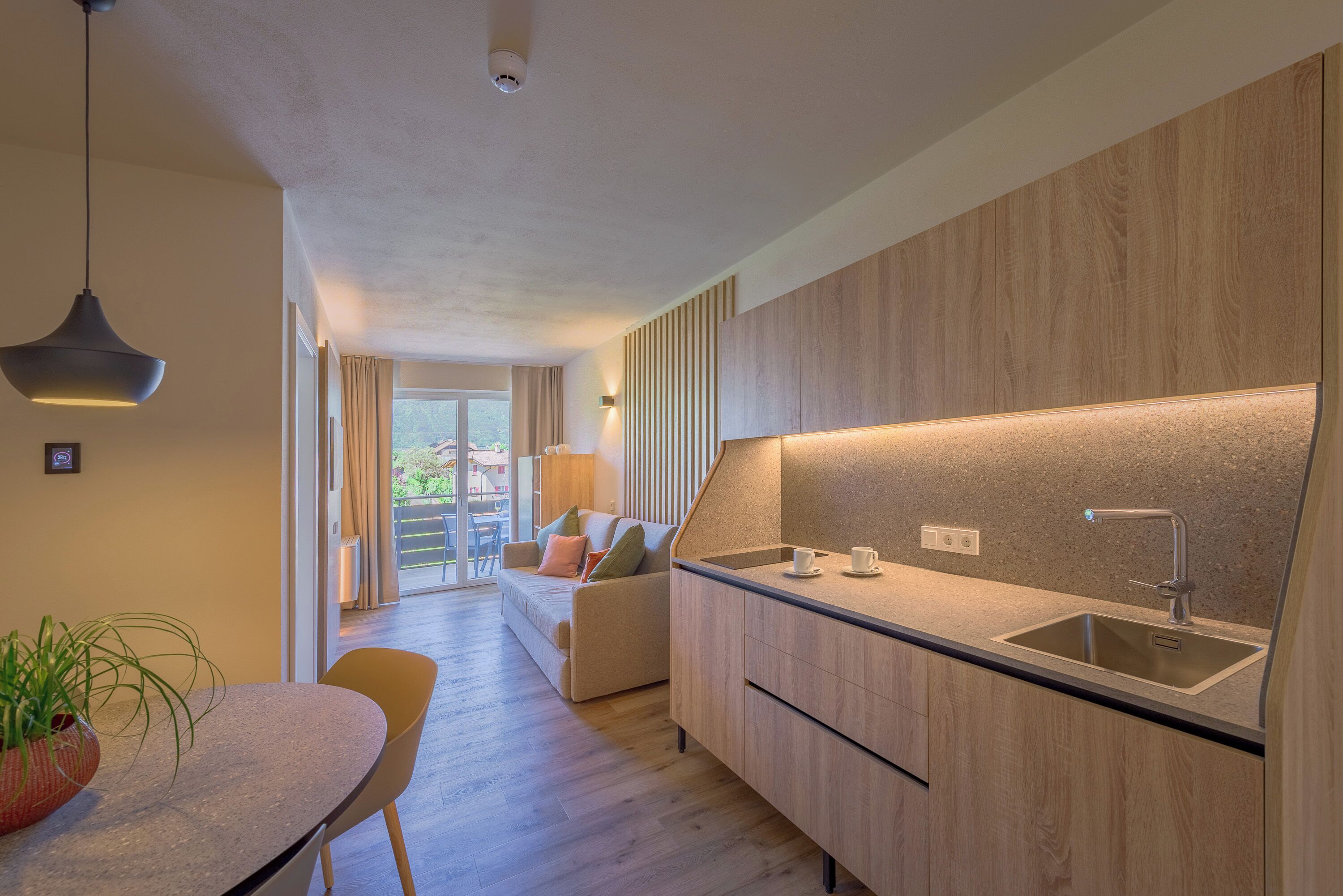 A modern kitchen with a built-in sink, a dining area with a table and chairs, and a living room with a sofa and a view of the outdoors.