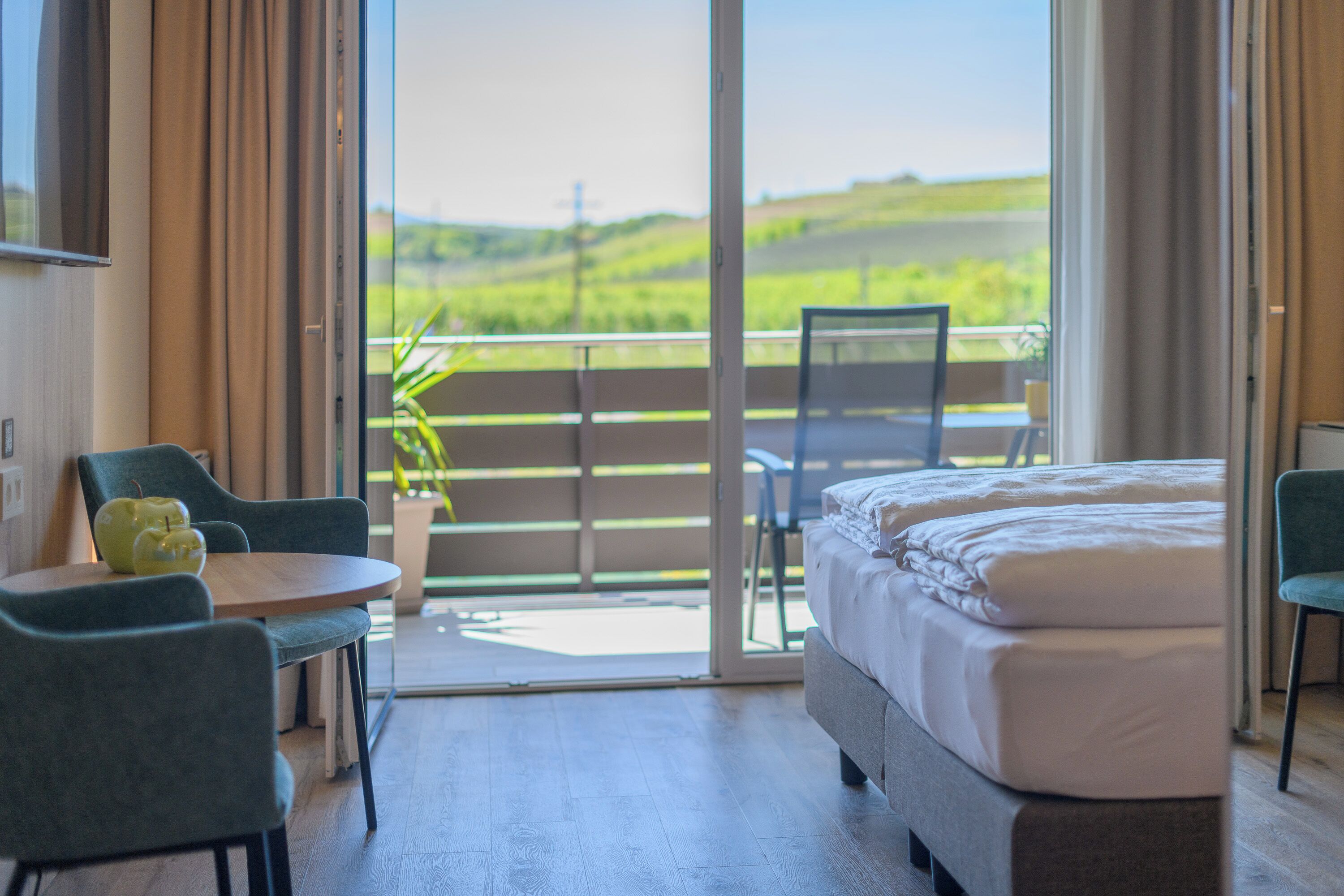 A hotel room with a bed, a table with chairs, a potted plant, and a balcony with a view of greenery.
