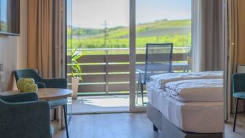 A hotel room with a bed, a table with chairs, a potted plant, and a balcony with a view of greenery.