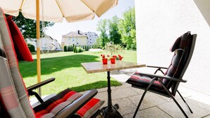 An outdoor patio with a table and chairs, a large umbrella, and a view of a green lawn and buildings in the background. - Haus am Mühlenweg (Bad Woerishofen)