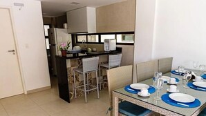 A dining area with a glass table, chairs, and a kitchen in the background. - Beira Mar C Piscina e Area Gourmet 113D (Ipojuca)