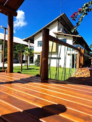 A wooden deck with a view of a two-story building with a white facade and a sloped roof. - Beira Mar C Piscina e Area Gourmet 113D (Ipojuca)
