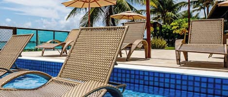 Un área junto a la piscina con sillas de descanso y sombrillas, rodeada de palmeras.