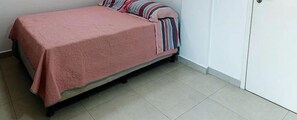 A single bed with a pink bedspread and striped pillows in a room with white walls and a tiled floor. - Beira Mar C Piscina e Area Gourmet 113D (Ipojuca)