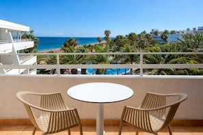 A balcony with two wicker chairs and a small round table, overlooking a beach with palm trees and a clear blue sky. - Sol Lanzarote All Inclusive (Puerto del Carmen)