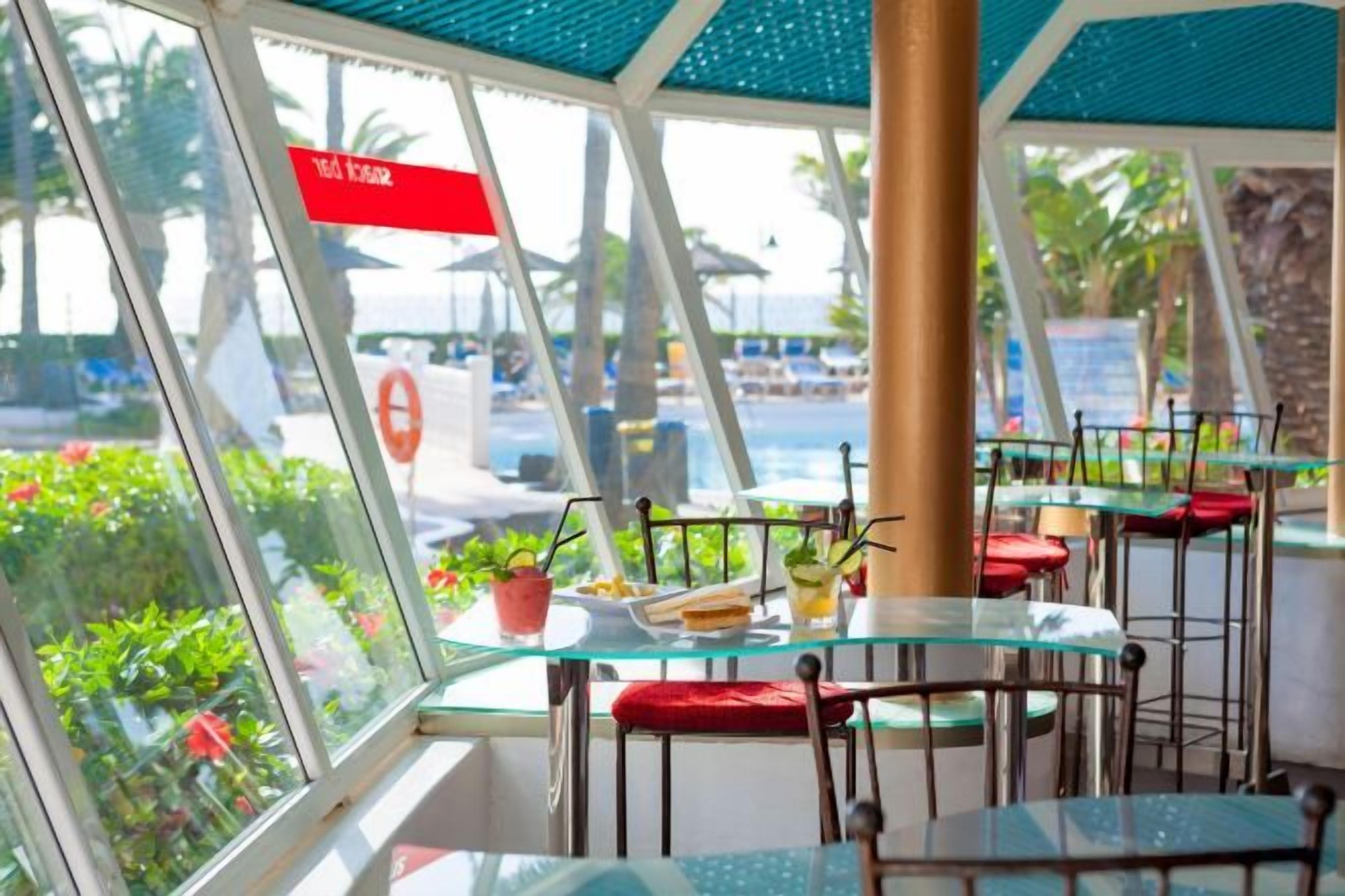Comedor con paredes de vidrio que cuenta con mesas y sillas, vista a una piscina y plantas tropicales.