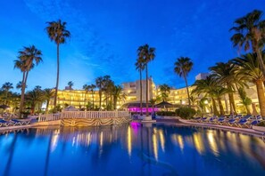 A hotel pool area with palm trees, lounge chairs, and a building with illuminated windows. - Sol Lanzarote All Inclusive (Puerto del Carmen)
