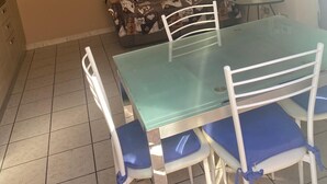 Ein Essbereich mit einem Tisch mit Glasplatte, weißen Stühlen mit blauen Kissen und einem gemusterten Sofa vor einer Wand, auf der sich ein kleiner Tisch und ein weißes Objekt befinden.