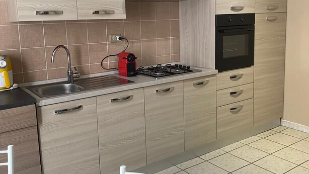 Eine moderne Küche mit hellen Holzschränken, einer Edelstahl-Dunstabzugshaube, einem Gasherd und einem eingebauten Backofen. Ein Glastisch mit einem Stuhl ist vorhanden, und ein weißer Stuhl ist im Vordergrund sichtbar.
