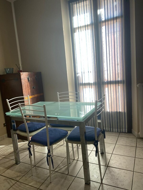 Un coin repas comprenant une table avec un plateau en verre et quatre chaises, un meuble en bois et une fenêtre équipée de stores verticaux.