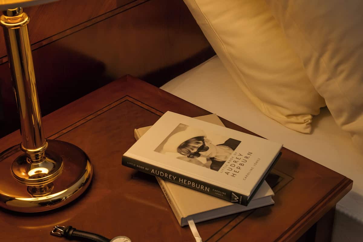 Una mesita de noche de madera con una lámpara, un libro titulado 'Little Book of Audrey Hepburn', un reloj de pulsera y un almohadón sobre una cama.