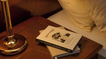 Une table de chevet en bois avec une lampe, un livre intitulé « Petits secrets d’Audrey Hepburn », une montre et un oreiller sur un lit.