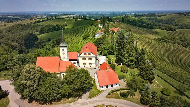 A large estate with a prominent building featuring a red roof and a tower, surrounded by lush green fields and a winding road. - Dvorec Jeruzalem (Ljutomer)