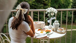 A woman sitting at a white metal table with a white tablecloth, eating breakfast. The table has a white vase with flowers, a white teapot, and a white cup. The woman is wearing a white top and a beige skirt. The background shows a green garden with trees and a white fence. - Dvorec Jeruzalem (Ljutomer)