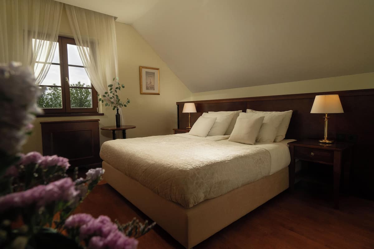Una cama bien hecha con cabecera de madera, una mesita de noche con lámpara y una ventana con cortinas traslúcidas.