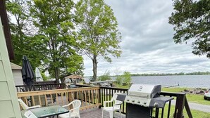 Eine Terrasse mit Grill, weißen Kunststoffstühlen und einem Tisch mit Blick auf einen See.