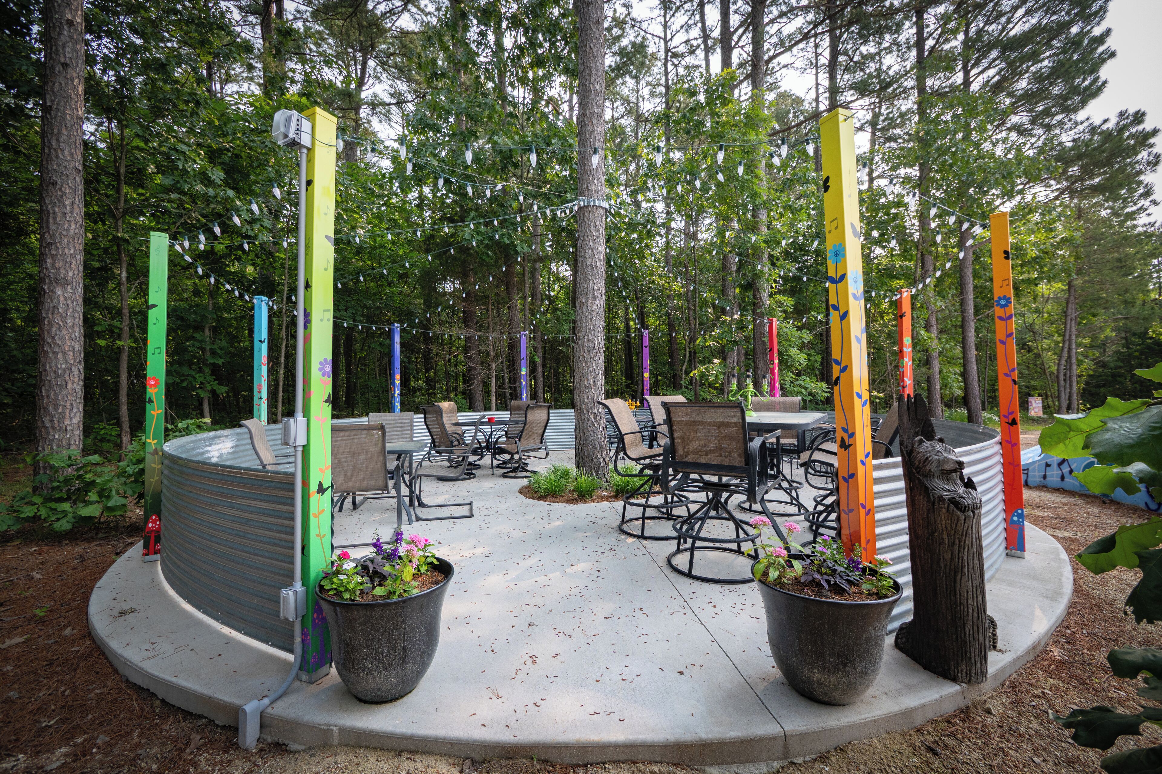 An outdoor patio area with a round table and chairs, surrounded by potted plants and colorful poles.
