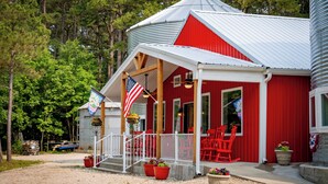A red barn-style building with a white porch, red chairs, and an American flag. - Sleeps 6 in a 2-story grain bins 2 queen beds 1 queen bunk & 2 full bathrooms. (Hermann)