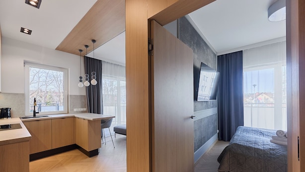 A modern hotel room with a wooden floor, a bed with a quilted blanket, a desk with a chair, a window with curtains, and a TV mounted on the wall. - Rezydencja Willora Świnoujście (Swinoujscie)