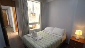 A neatly made bed with a striped bedspread and white pillows, a bedside lamp, and a view of a hallway and another room through a window. - Apartment 5 minutes from downtown (Paracas)