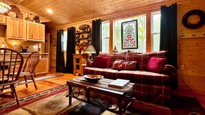 L’intérieur d’un chalet accueillant, avec un plafond en bois, un canapé agrémenté de coussins à carreaux, une table à manger avec des chaises et une fenêtre ornée de vitraux.