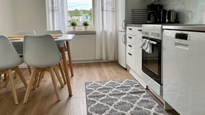 A modern kitchen with white cabinets, a microwave, a coffee maker, and a dishwasher. There is a dining area with a table and chairs, and a window with curtains.