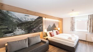 Ein modernes Hotelzimmer mit einem großen Bett, einer Couch, einem Wandbild einer Berglandschaft und einem Fenster mit Vorhängen.