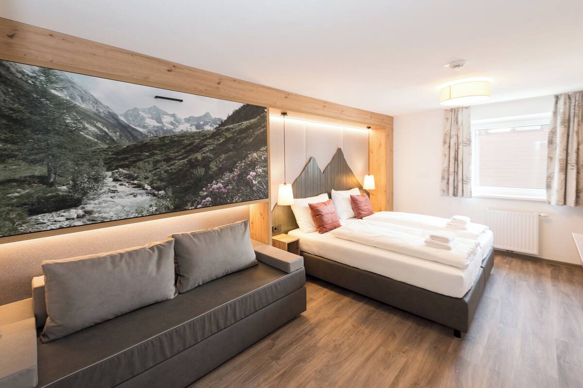 A modern hotel room with a large bed, a sofa, a mounted artwork of a mountainous landscape, and a window with curtains.