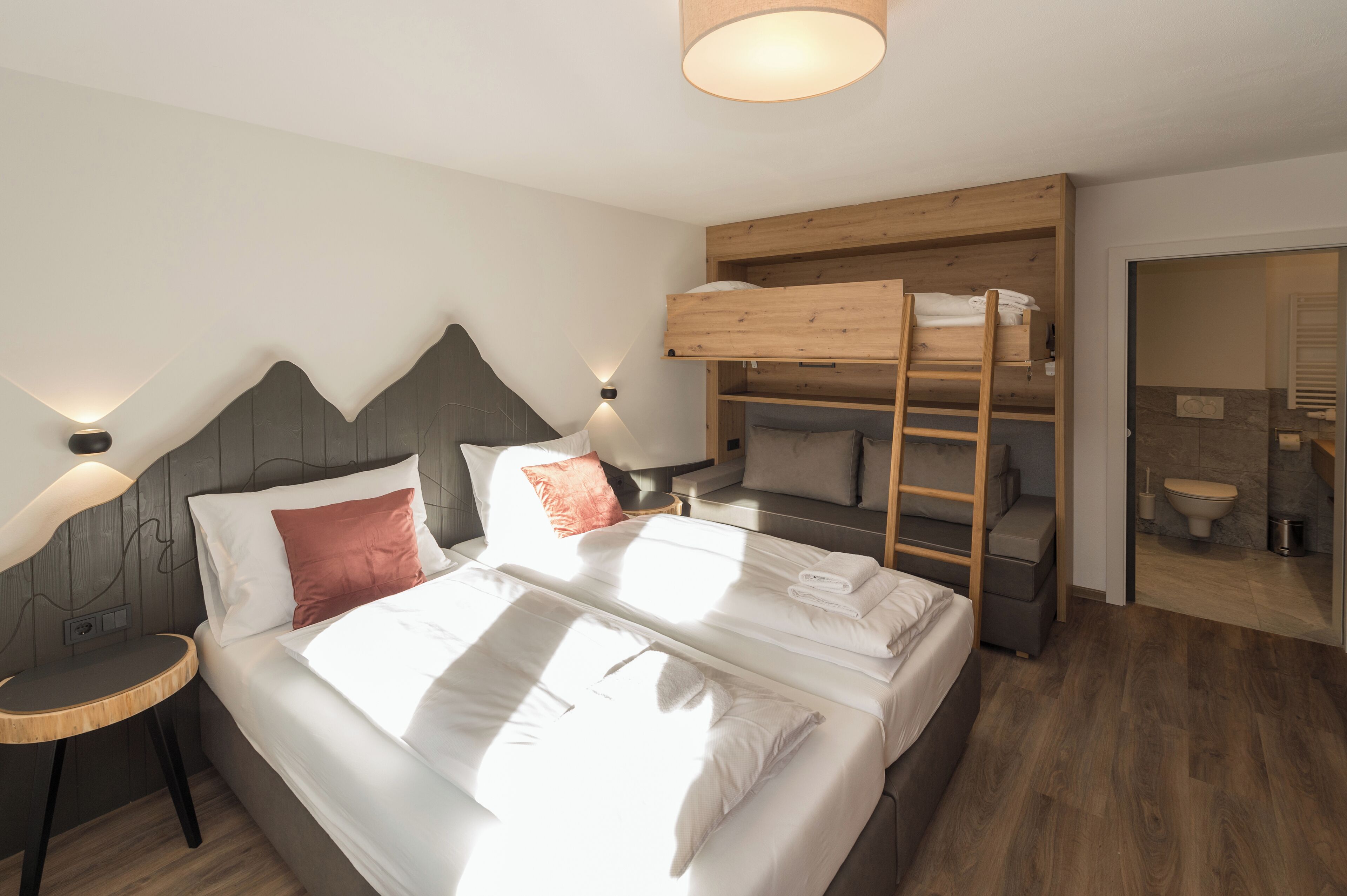 Quarto de hotel moderno com beliche, uma cama separada, uma mesinha de cabeceira e um banheiro visível através de uma porta aberta.
