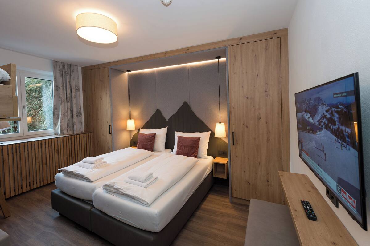 A modern hotel room with a large bed, wooden paneling, a television, and a window with a view of trees.