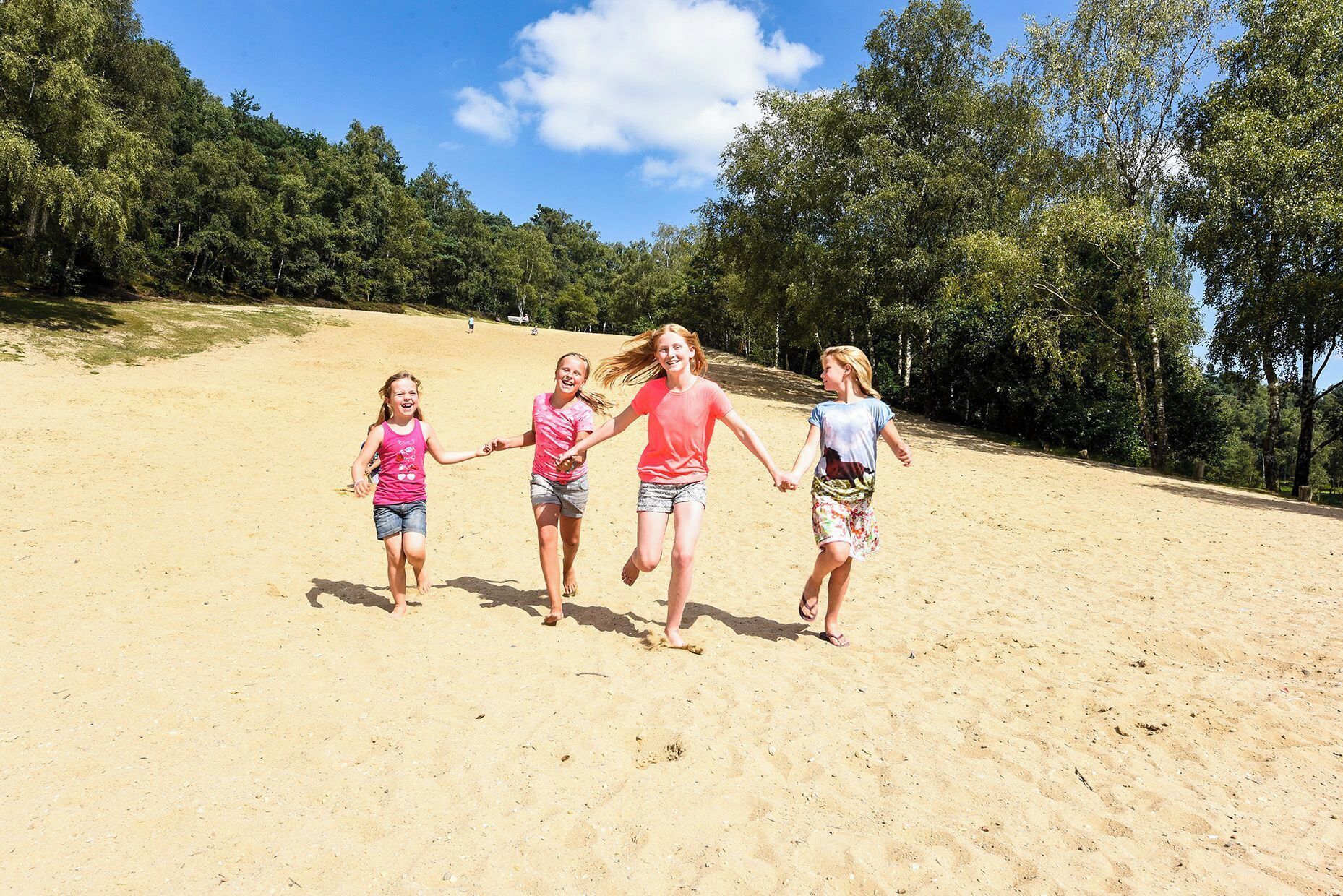 Four individuals running on a sandy beach with trees in the background.