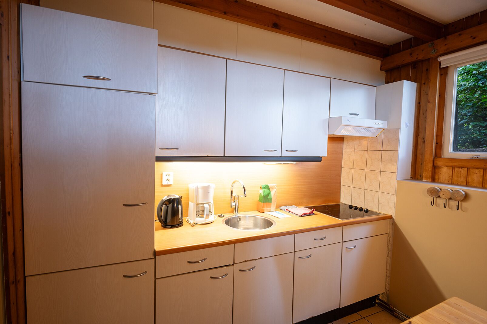 A compact kitchen with white cabinets, a wooden countertop, a stainless steel sink, a coffee maker, and a kettle.