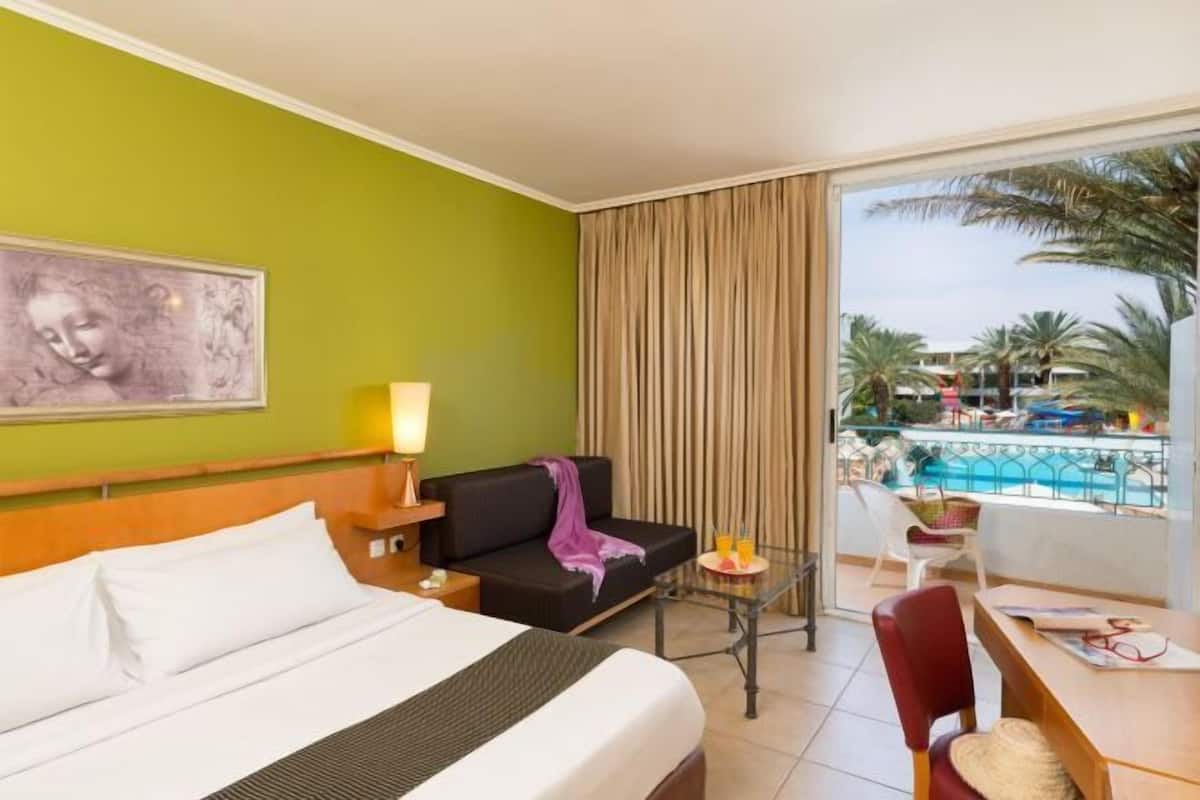 Une chambre d’hôtel avec un lit, une petite table avec des boissons, une chaise, un canapé et une vue sur une piscine et des palmiers.