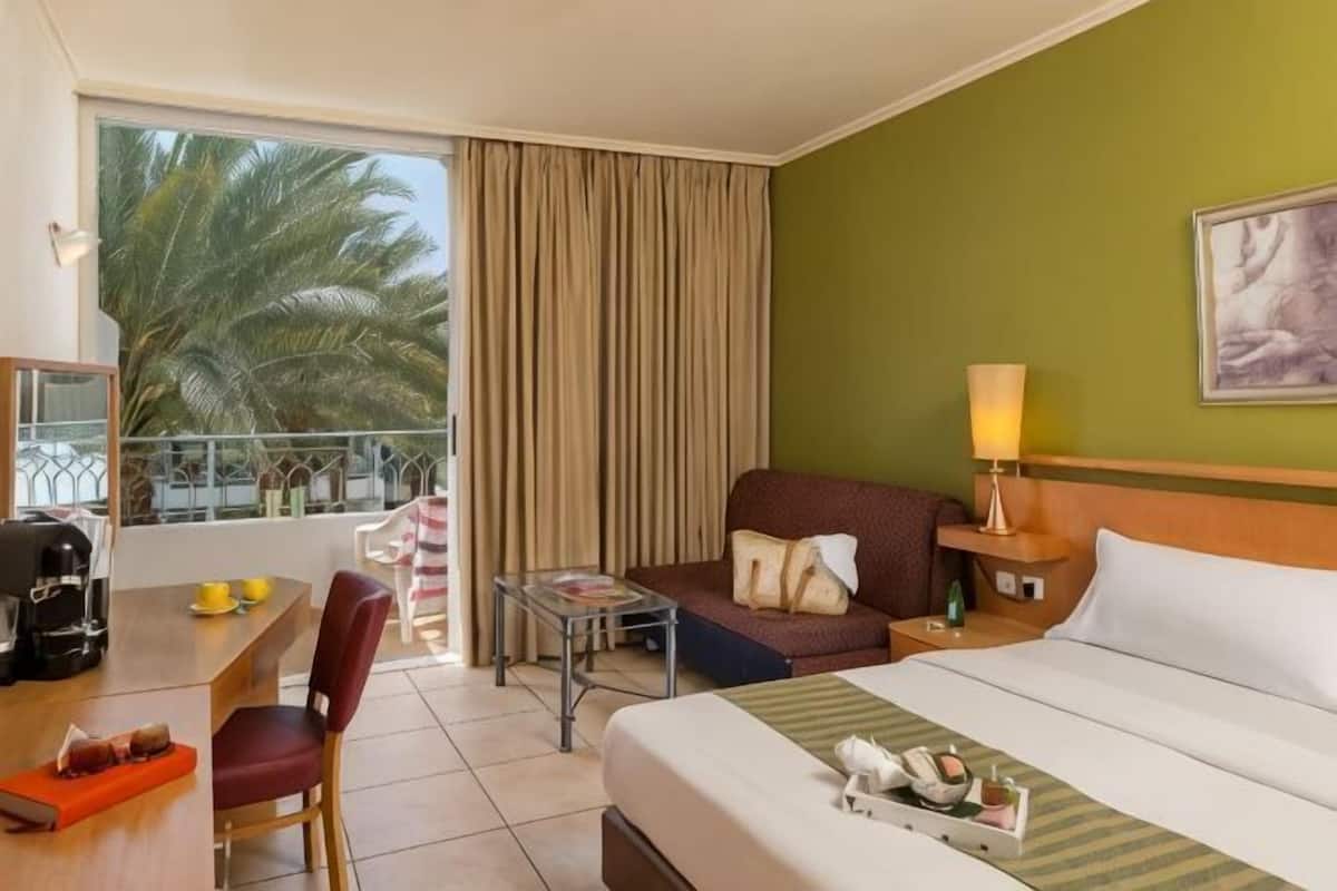 Une chambre d’hôtel comprenant un lit, un bureau avec une chaise, une petite table, un canapé et un balcon avec des rideaux.