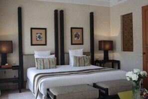 Una habitación de hotel con una cama grande, dos mesitas de noche con lámparas, un banco y un jarrón con flores.