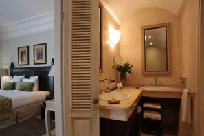 Habitación de hotel con cama, baño con lavamanos y espejo, y una puerta que da a otra habitación.
