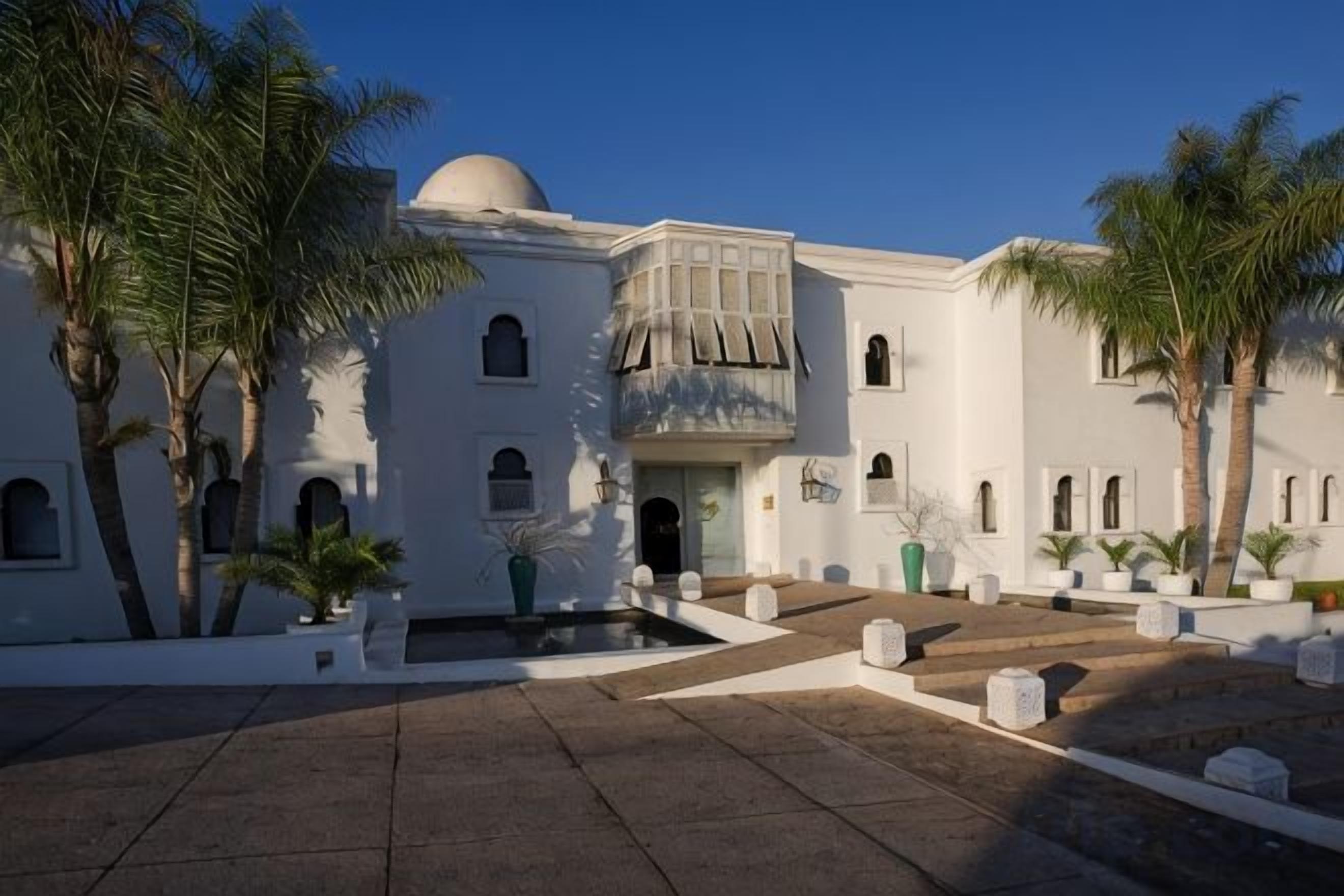 A white building with a dome, multiple windows, and a courtyard with palm trees.