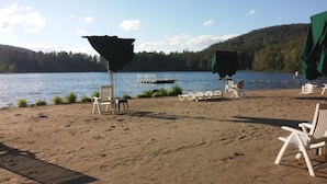 Une plage avec des chaises longues blanches et des parasols, une jetée avec un bateau, et une colline boisée en arrière-plan.