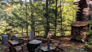 Un espace extérieur aménagé dans un style rustique, avec des chaises en bois et un foyer, le tout entouré d’une forêt dense.