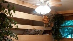 Une chambre confortable, aménagée dans le style d’un chalet, avec un lit, une table de chevet, un ventilateur de plafond et une fenêtre donnant sur la forêt.