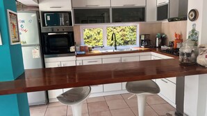 Una cocina con armarios blancos, un microondas, un horno, un fregadero y un ventanal con vistas a zonas verdes. Hay dos taburetes de bar y una cafetera.