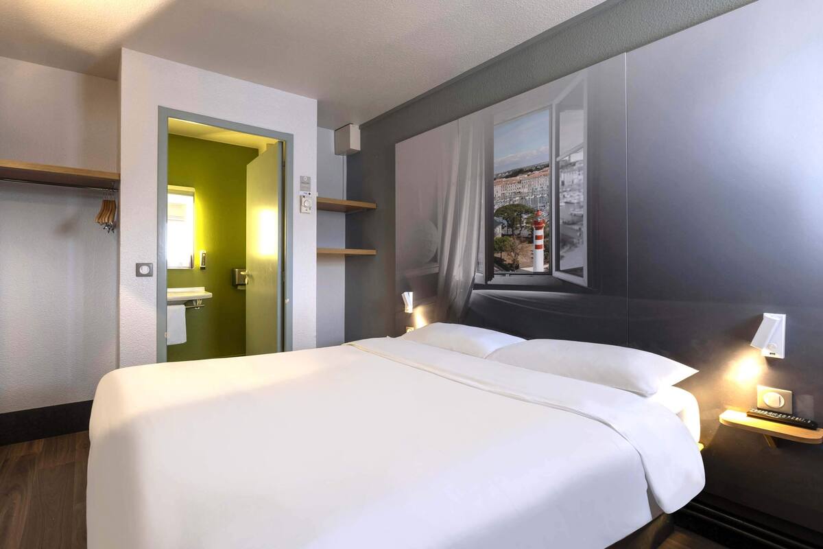 A hotel room with a large bed, a window with a view of buildings, a door leading to a bathroom, and a bedside table with a lamp.